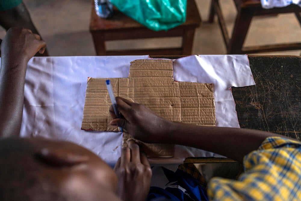 Over a desk a young girl is tracing the silhouette of a cardboard cross over cotton fabric, beginning the process to make a home made menstrual pad.